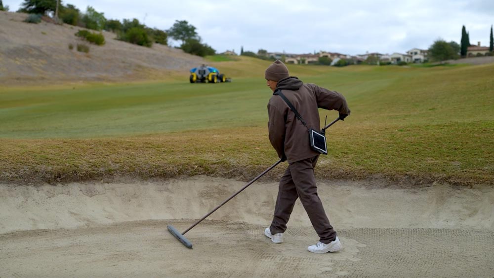 A man raking a golf sand bunker with a robotic mower working in the background